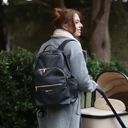 Woman walking with a baby stroller and carrying a black changing backpack outdoors.