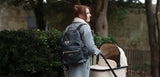 Woman walking with a baby stroller and carrying a black changing backpack outdoors.
