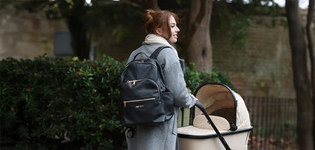 Woman walking with a baby stroller and carrying a black changing backpack outdoors.