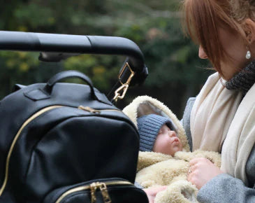 Woman holding a baby with a changing backpack hanging from the stroller with a green blurred background