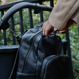 Black leather look changing backpack hanging from a stroller showing how it can be unzipped. 